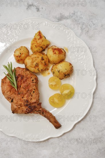 Delicious roasted pork rib roast is plated next to golden-brown crispy potatoes and tomato cherry. The meal is garnished with a sprig of rosemary, enhancing its savory appeal