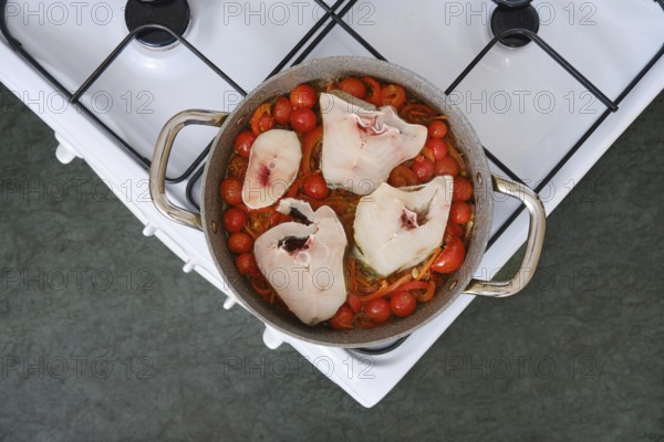Bright red tomatoes and colorful peppers surround large pieces of cod fillet in a pot on a stove