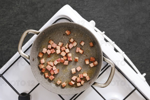 Diced smoked bacon is sizzling in a pan on a stove, browning evenly in oil. The dark textured background creates a contrasting backdrop, highlighting the cooking process for a delicious meal