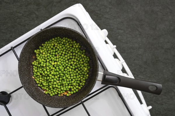 Fresh green peas are being cooked in a black pot on a white stovetop. Steam rises gently, indicating heat is applied
