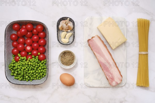 Overhead view of fresh cherry tomatoes, green peas, smoked bacon, garlic, parmesan cheese, spaghetti and an egg on a light kitchen counter