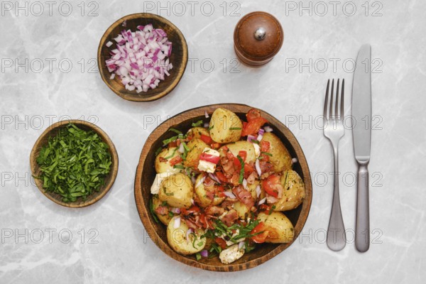 Greek potato salad in a wooden bowl, topped with tomatoes, onions, feta cheese and herbs. Additional bowls with chopped onions and cilantro accompany it, set on a marble table