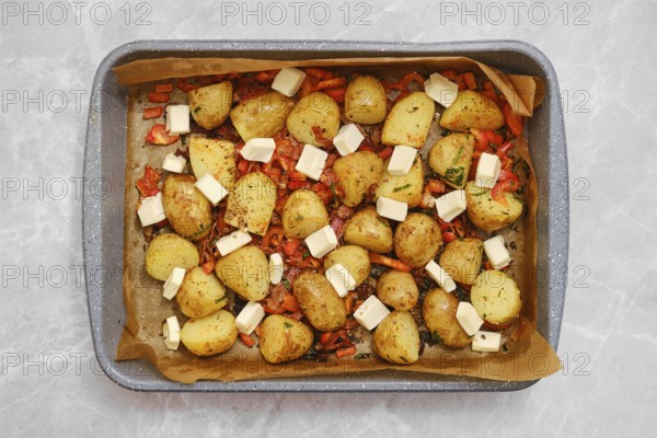 Golden roasted potatoes are arranged with pieces of feta cheese and herbs on a parchment in a baking tray. Colorful roasted vegetables accompany them, creating a delightful, mouthwatering dish
