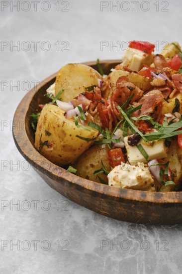 A bowl filled with cooked potatoes mixed with fresh vegetables, herbs, bacon and cheese. The dish is garnished with red onions and chopped parsley