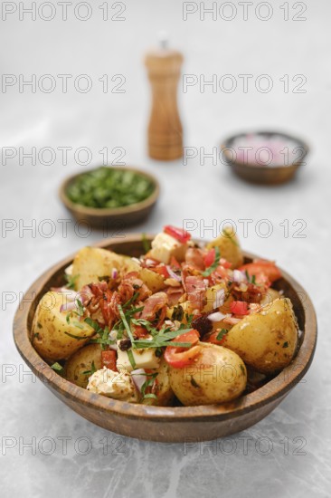 A delicious bowl of roasted new potatoes is garnished with bacon, fresh herbs, chopped onions, and colorful vegetables, creating a vibrant dish perfect for sharing at a family gathering or picnic