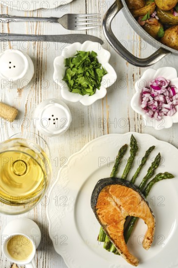 Salmon steak on a bed of asparagus, accompanied by fresh herbs and onions. A glass of white wine and a bowl of potatoes complete the enticing meal setting