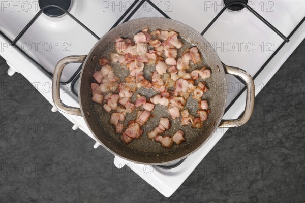Bacon strips are sizzling and browning in a stainless steel frying pan placed on a stovetop. The kitchen setting is bright and clean, indicating a typical morning meal preparation