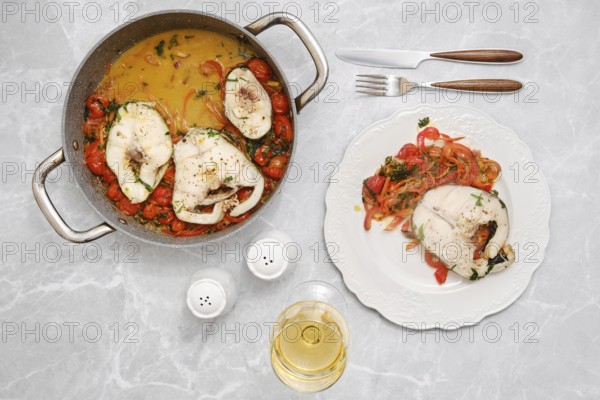 A flavorful fish dish is being prepared in a pot alongside a vibrant mix of tomatoes, carrots and herbs. The meal is elegantly plated with vegetables and served with a glass of white wine