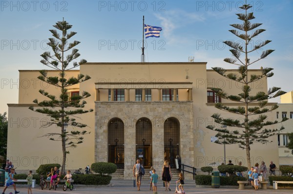 Large symmetrical building with Greek flag surrounded by people in the evening light, Kos Town, Kos, Dodecanese, Greek Islands, Greece