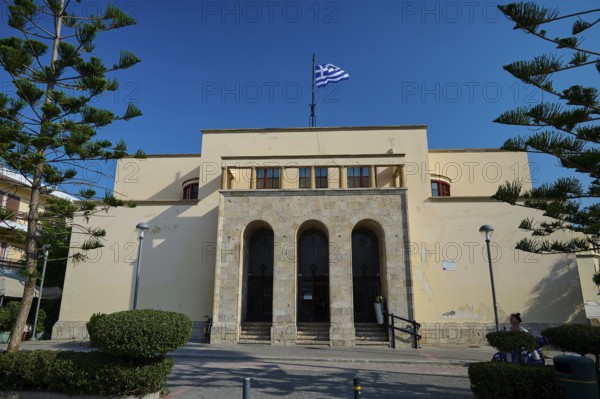 Archaeological Museum, beige building with Greek flag on the roof, symmetrical architecture, surrounded by trees under a clear sky, Kos Town, Kos, Dodecanese, Greek Islands, Greece