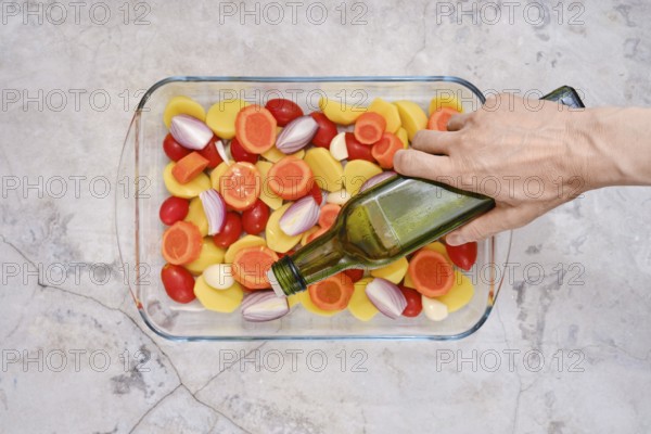 Pouring olive oil on slices of fresh vegetables including tomatoes, carrots, potatoes and onions ready for roasting