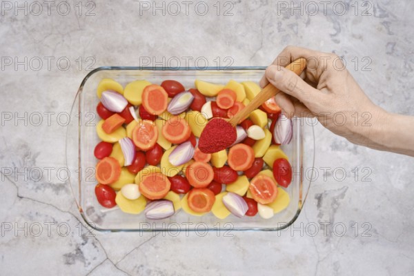 A hand holds a wooden spoon with smoked paprika above a glass dish filled with vibrant vegetables including bell peppers, tomatoes, and shallots, ready to be seasoned for a delicious meal