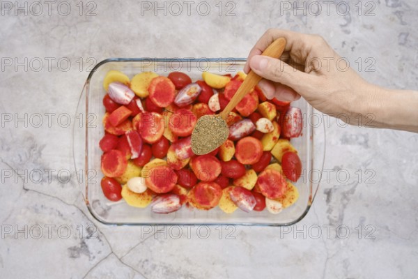 A hand sprinkles oregano over a colorful mixture of tomatoes and other vegetables in a glass baking dish. This preparation is a step for baking turkey breast for a flavorful meal