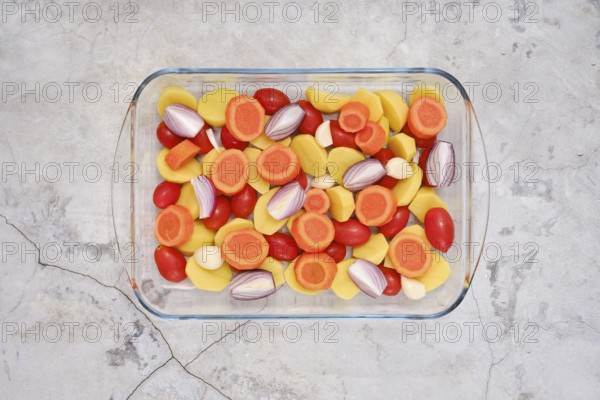 Fresh colourful vegetables like carrots, tomatoes, potatoes, onion and garlic cloves all neatly arranged in a glass baking dish