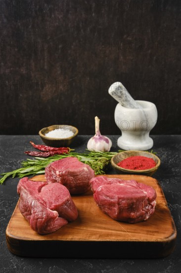 Several cuts of fresh beef tenderloin are arranged on a wooden cutting board, surrounded by spices like paprika powder and salt. Chopped herbs and garlic add color to the kitchen setup, ready for meal preparation