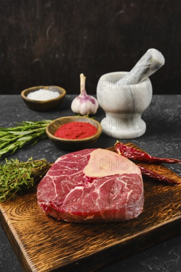 A fresh cut of beef rests on a wooden board, surrounded by garlic, herbs, and various spices. A marble mortar and pestle sit nearby, ready for seasoning