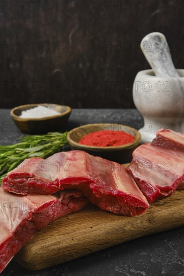 A selection of raw beef ribs is arranged on a wooden cutting board alongside bowls of seasoning. Salt and red seasoning are visible, while fresh herbs add a touch of color and flavor