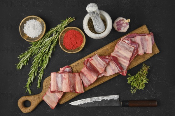 Fresh raw beef short ribs lay on a wooden cutting board alongside bowls of seasoning, herbs, and a knife, indicating a cooking preparation in a home kitchen for a flavorful meal