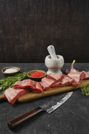 Raw beef ribs are on a wooden cutting board alongside a mortar and pestle. Herbs and spices are arranged nearby, creating an inviting atmosphere for cooking in a rustic kitchen