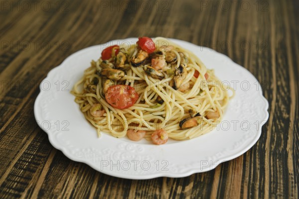 A delicious plate of spaghetti topped with shrimp and mussels, complemented by fresh cherry tomatoes and herbs, sits on a beautifully designed white plate against a rustic wooden table background