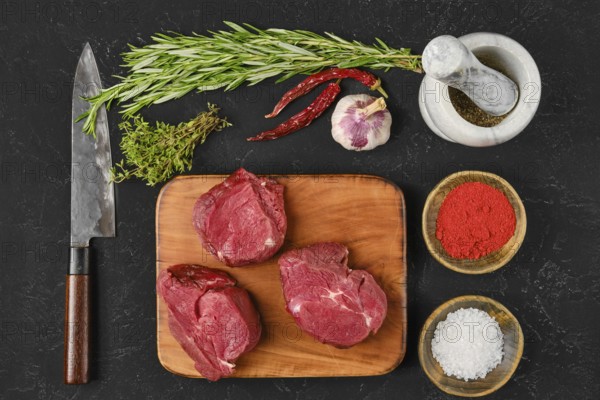 Fresh cuts of beef filet mignon are arranged on a wooden cutting board, surrounded by herbs, spices, and a knife. Ingredients like garlic and chili add aroma, perfect for cooking flavorful meals