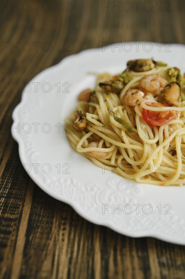 A plate of seafood spaghetti features fresh pasta topped with shrimp, mussels, and vegetables. It is elegantly arranged on a white decorative plate, against a warm wooden table