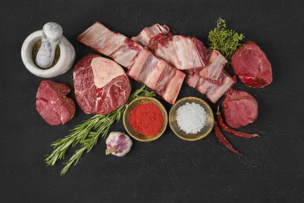 A variety of fresh cuts of beef are arranged on a dark surface, surrounded by herbs, spices, and garlic, showcasing the ingredients needed for a flavorful meal preparation