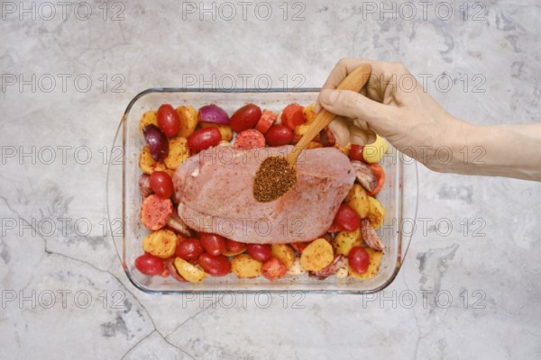 A hand sprinkles a blend of spices over a raw turkey breast resting on a bed of vibrant red and yellow potatoes in a glass baking dish, ready for a flavorful cooking process