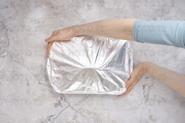 A hand holds a baking dish covered with aluminum foil, ready for the oven. The setting features a gray stone surface, indicating the beginning of a cooking process for a turkey breast