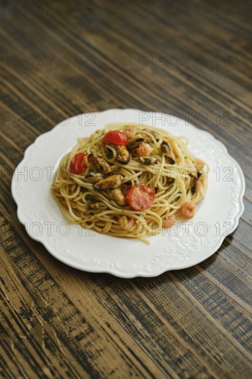 A serving of spaghetti topped with fresh seafood and cherry tomatoes is showcased on a white plate atop a rustic wooden table. This dish highlights vibrant colors and appetizing textures