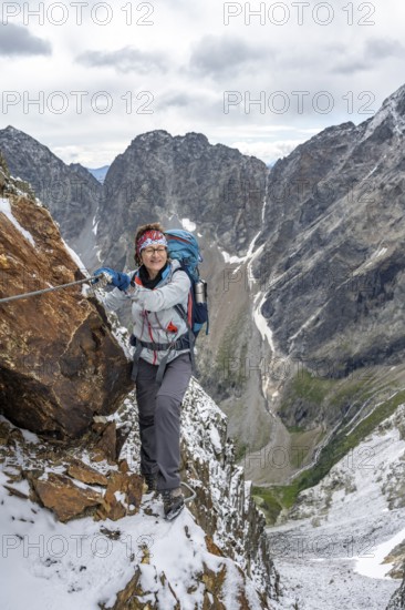 Mountaineer on a rope-secured path in a steep rocky mountain landscape with fresh snow in summer, behind mountain peaks Großer Friedrichskopf and Georgskopf, ascent to Hornscharte, Schobergruppe, Hohe Tauern National Park, Carinthia, Austria