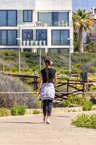 Young woman walking outdoors on a footpath in a residential area and listening to music with headphones from behind, Mallorca, Spain