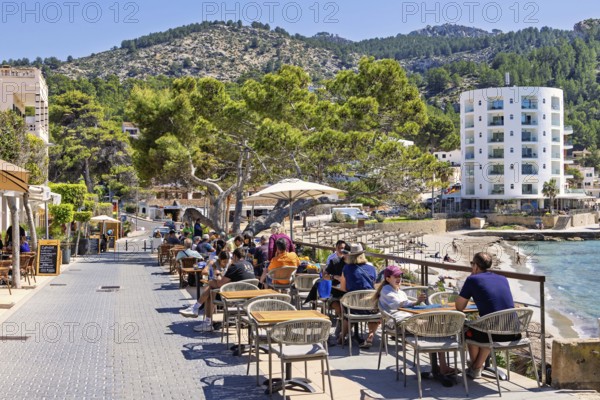 Tourists sitting in an outdoor restaurant on a boardwalk by the Mediterranean Sea at a tourist resort a sunny summer day, Sant Elm, Mallorca, Spain