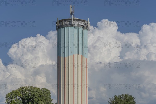 A tall water tower, painted with blue and white stripes, sits enthroned in a green landscape. Fluffy white clouds fill the bright blue sky, creating a picturesque scene during daylight hours. Bas rhin, Alsace, grand est, France