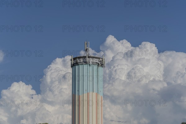 A tall water tower, painted in bright colours, rises against a backdrop of flowing white clouds. The clear blue sky enhances the vibrant colours of this building in a peaceful suburban landscape. Bas rhin, Alsace, grand est, France