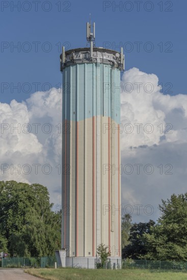 A tall water tower, painted with blue and white stripes, sits enthroned in a green landscape. Fluffy white clouds fill the bright blue sky, creating a picturesque scene during daylight hours. Bas rhin, Alsace, grand est, France