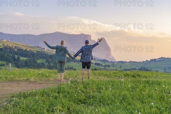 A couple raises their arms in joy at the sight of the mountain landscape and the evening sky, Alpe di Siusi, Dolomites, South Tyrol, Italy