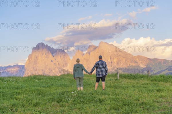 A couple stands hand in hand in a meadow in front of an impressive mountain landscape, Alpe di Siusi, Dolomites, South Tyrol, Italy