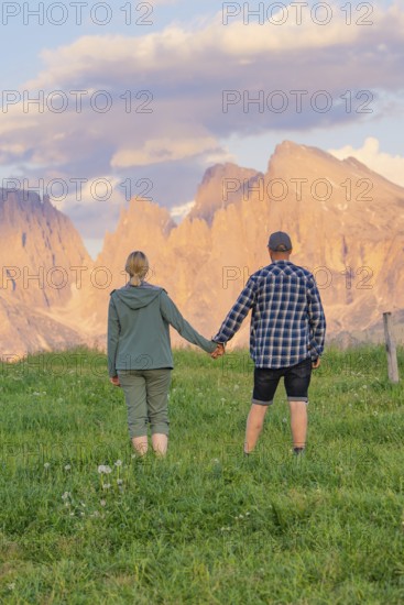 A couple holding hands in a meadow against an impressive mountain backdrop, Alpe di Siusi, Dolomites, South Tyrol, Italy