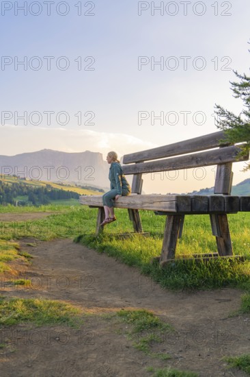 A woman sits on a large bench with a view of the tranquil mountain landscape, Alpe di Siusi, Dolomites, South Tyrol, Italy