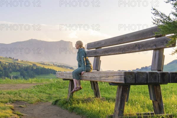A woman sits on an oversized bench and gazes at the picturesque mountain landscape, Alpe di Siusi, Dolomites, South Tyrol, Italy