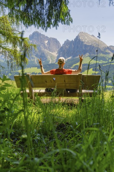 Person on a bench showing a peace sign in front of a mountain landscape, Alpe di Siusi, Dolomites, South Tyrol, Italy