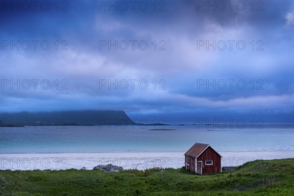 A red wooden hut on the sandy beach of Ramberg (Rambergstranda), sea and mountains. At night at the time of the midnight sun. Clouds in the sky. Early summer. Ramberg, Flakstadoya, Lofoten, Norway