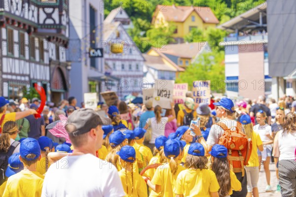 Children in yellow shirts and blue caps march with signs through a busy half-timbered street, 950 years of Calw, Calw parade, Black Forest, Germany