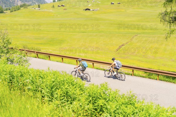Two cyclists on a road leading through a vast green landscape, Alpe di Siusi, Dolomites, South Tyrol, Italy