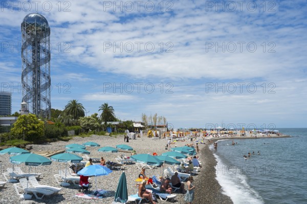 Beach with many visitors under umbrellas, a view of the sea and a distinctive tower in the background, Alphabetic Tower, Georgian Alphabet Tower, Miracle Park, Batumi, Black Sea, Ajara region, Adjara, Autonomous Republic, Georgia