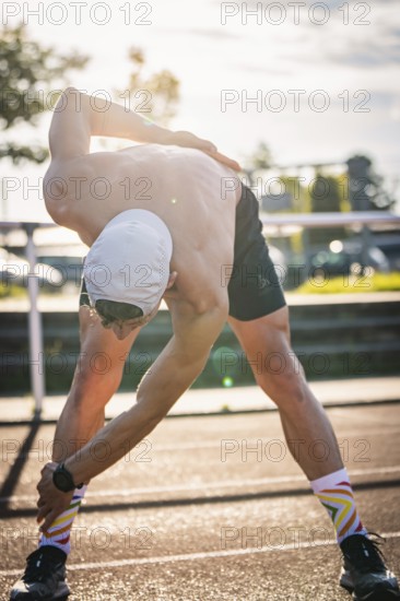 Man stretches intensively in sporty clothing in sunlight on a running track, triathlon, Calw, Germany