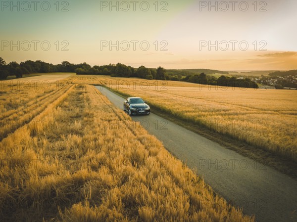 A car drives on a country road between golden fields at sunset, BMW i7, Deer E-Carsharing, Calw, Germany