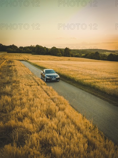 A car drives along a road surrounded by vast fields of corn with a colorful sunset in the background, BMW i7 electric car, deer e-car sharing, Calw, Germany
