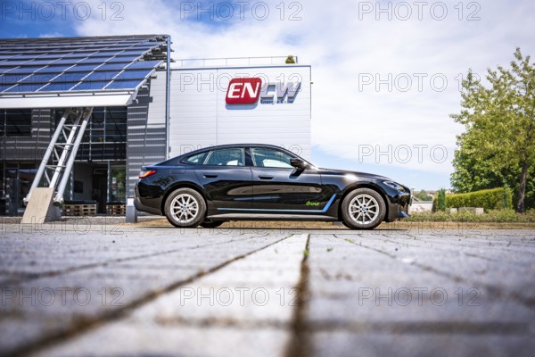 Black car on the side of a modern building with ENCW logo on a paved path, BMW i7 electric car, Deer e-Carsharing, Calw, Germany